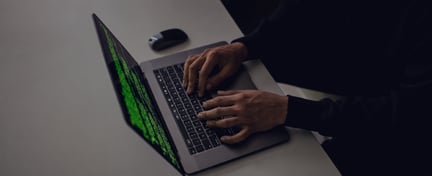 Person typing on a laptop displaying green code on a dark screen, viewed from an overhead angle in a minimalist workspace